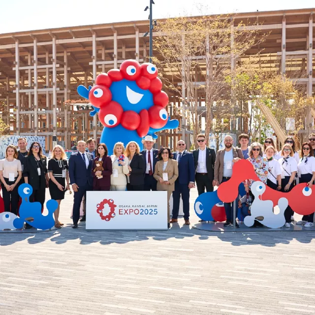 grupa osób stojących przed drewnianą konstrukcją na świeżym powietrzu. Z przodu duży baner z napisem 'EXPO 2025' oraz logo wydarzenia. Wokół nich ustawione są duże, kolorowe figury balonowe oraz kartonowe postacie w kolorach czerwonym i niebieskim, przypominające bajkowe stwory z dużymi oczami. 