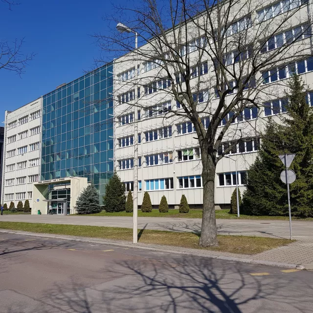 Dwa budynki stojace obok siebie, jeden w kolorze szarym, drugi przeszklony od parteru do ostatniego, 6, piętra w części znajdującej się nad wejściem